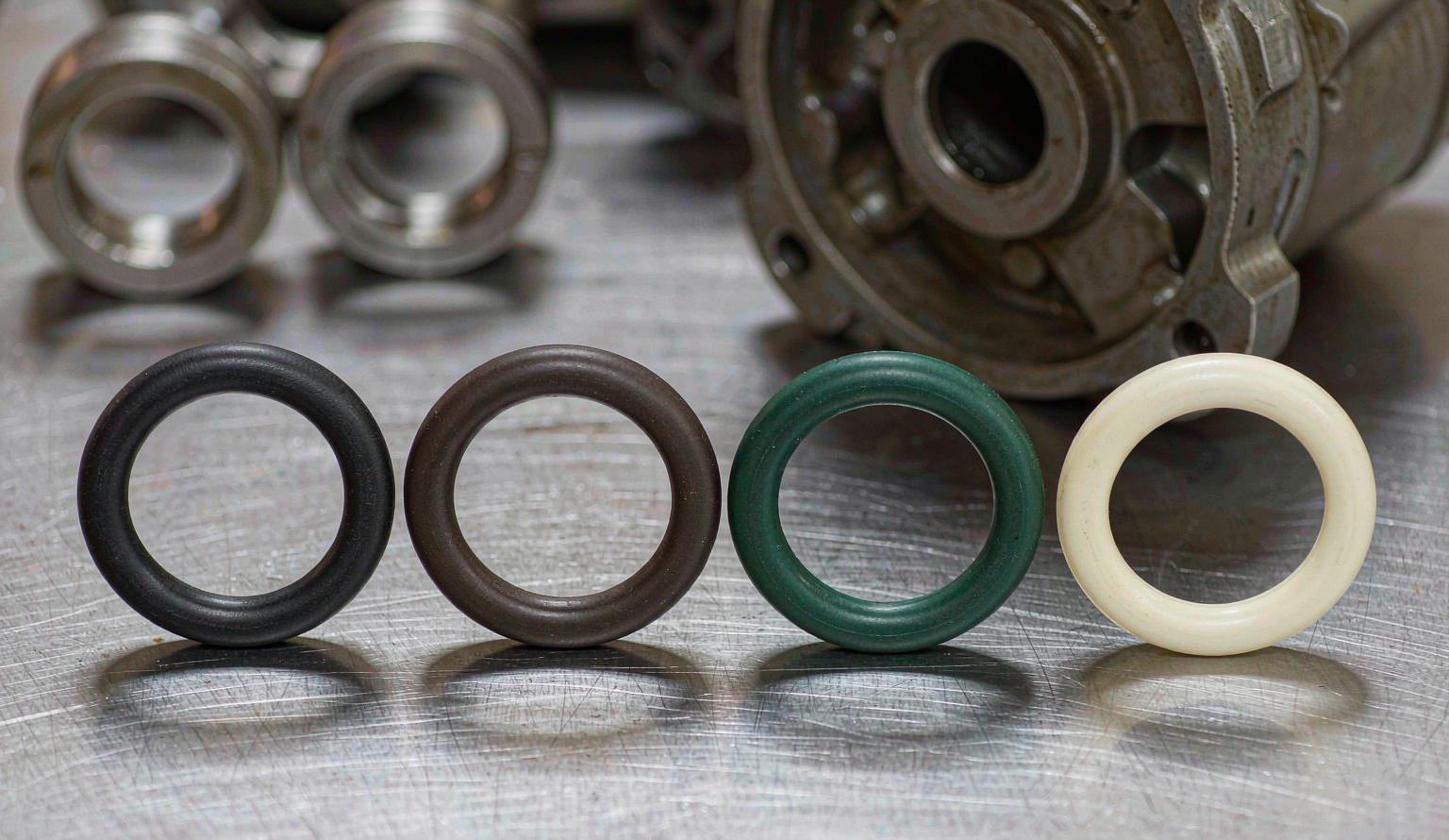 Four oilfield seal compounds compared side by side: black HNBR, brown FKM, green AFLAS and cream FFKM O-rings on a stainless steel workshop bench with machined protector hardware